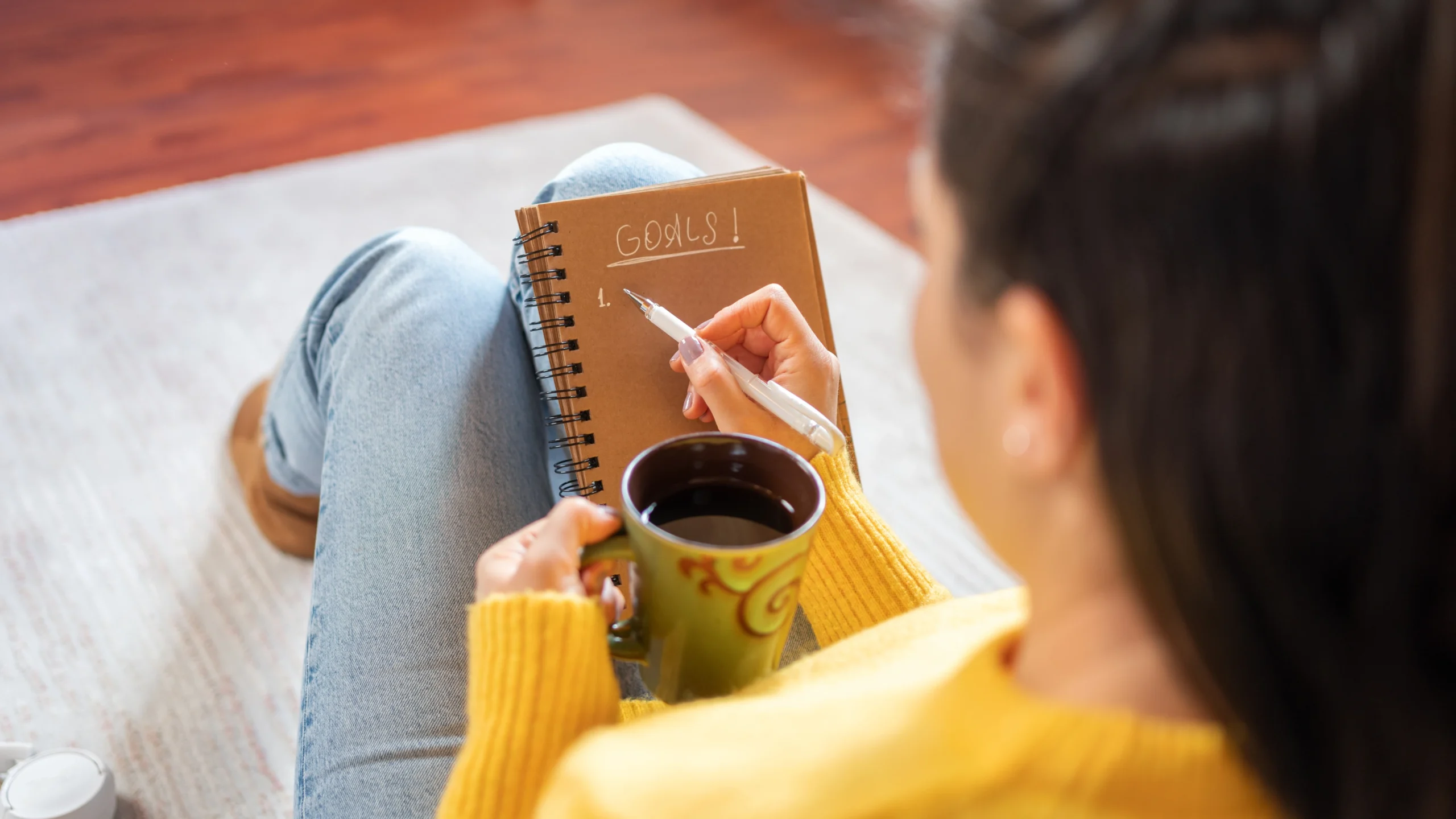 Woman writing marketing resolutions in a notebook while holding a coffee mug, planning for success in 2025.