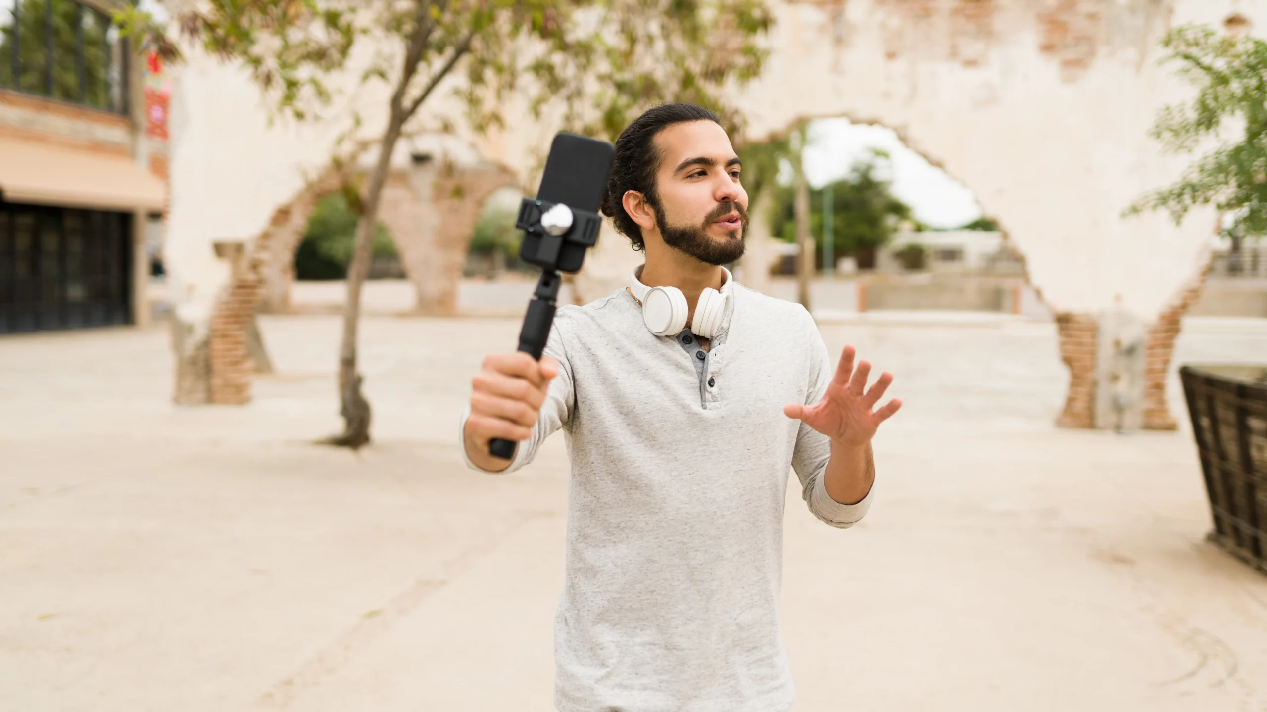 A young man recording a short-form video with a smartphone mounted on a stabilizer, wearing headphones around his neck, standing in an outdoor urban setting with historic architecture in the background. This image represents content creation and storytelling through short-form videos.